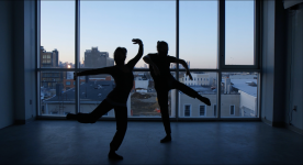 Two dancers in motion silhouetted against a window that looks out over a city skyline. Photo by Christian Pinto.